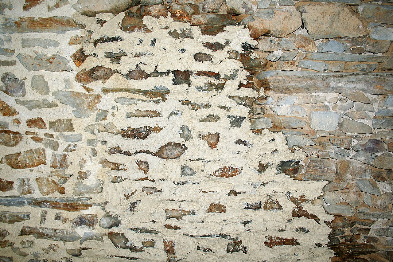 Mur ancien en pierre montrant des zones dégarnies, du mortier de chaux en cours d’application et un joint fini