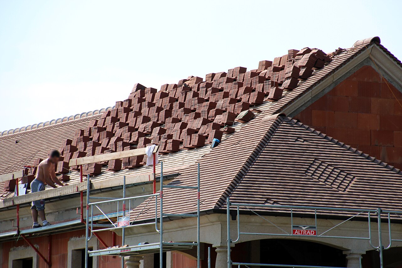 Pose de tuiles en terre cuite sur une toiture en construction, avec échafaudage et tuiles à répartir